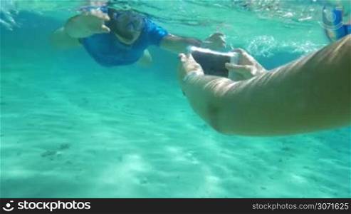 Slow motion of two people diving to get good underwater shot. Woman using smartphone in water-proof case to take photos or shoot video of a friendly man in snorkel