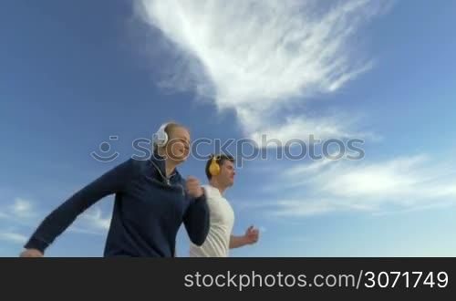 Slow motion of two runners during outdoor training. Happy couple jogging to music in headphones against background of blue cloudy sky