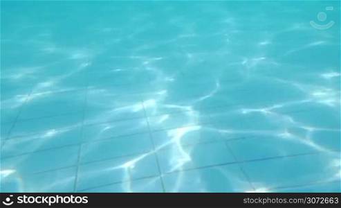 Slow motion of waving clear blue water in the swimming pool. It sparkling in the sun and creating shiny wavy pattern on the tiled floor