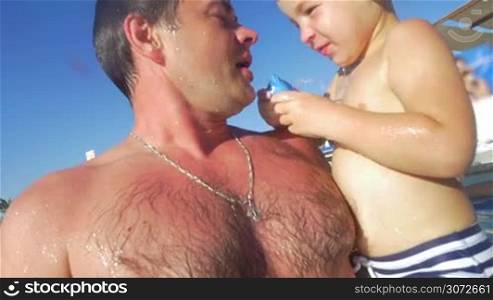 Slow motion of young father and little son having fun in the outdoor swimming pool. Boy squeezing water from the rubber into dads mouth and it making him laugh