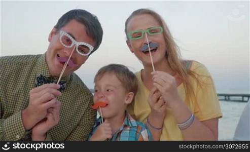 Slow motion of young happy family of three with paper hipster glasses and moustache outdoor. Parents and child having fun while making goofy faces. Modern trendy style