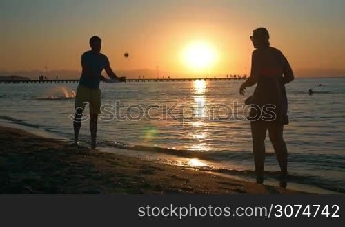 Slow motion of young man and woman playing tennis by the sea at sunset. Golden sun reflecting in dark water. Active and carefree summer vacation