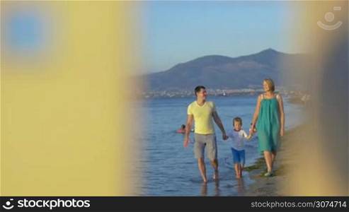 Slow motion of young parents with son walking along the sea, view through the door of house model with following focus on it. Family house, villa or vacation home concept