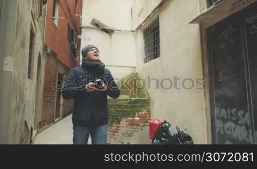 Slow motion of young tourist walking in the street of old Italy among worn buildings. He looking around and shooting video with retro camera