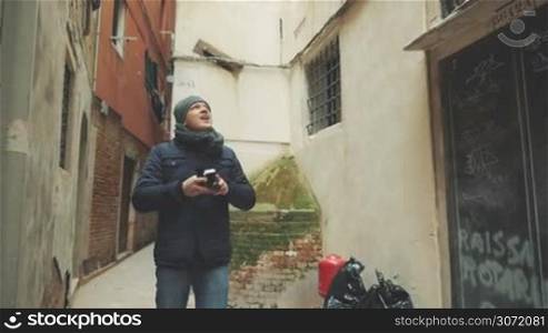 Slow motion of young tourist walking in the street of old Italy among worn buildings. He looking around and shooting video with retro camera