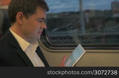 Slow motion shot of a man wearing business suit traveling by train. He is reading something funny in tablet and laughing.