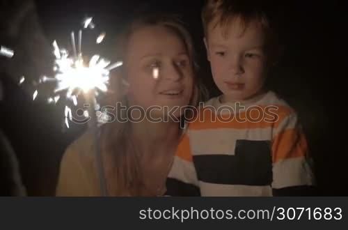 Slow motion shot of a young woman and her little brother looking at sparkles of Bengale fire. Woman is squatting near the boy and tell him something.