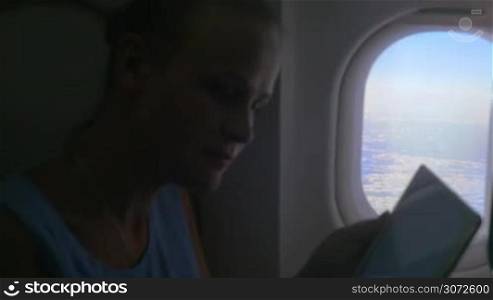 Slow motion shot of a young woman sitting on the plane and typing in tablet PC, then she&acute;s closing the blind on the window.