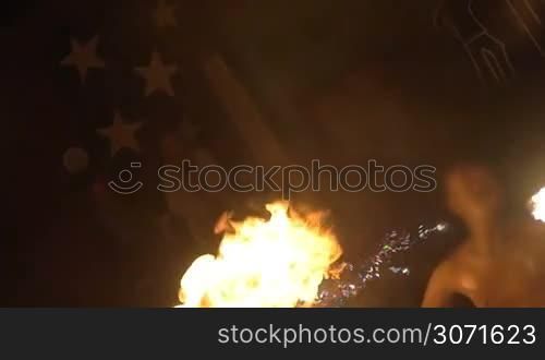 Slow motion shot of an episode of fire show. Dark-skinned artist blows fire with two torches in turns.