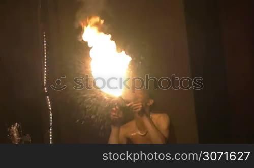 Slow motion shot of an episode of fire show. Dark-skinned artist breathe fire in the night.