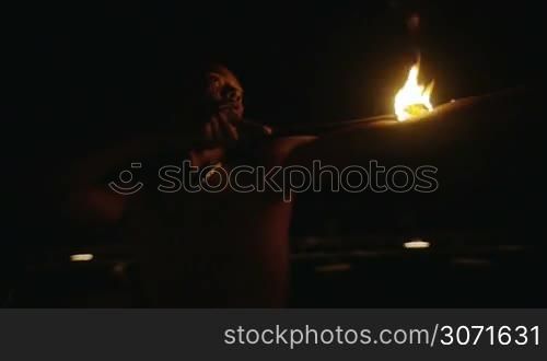 Slow motion shot of an episode of fire show. Dark-skinned artist puts burning torch on his arm and body.