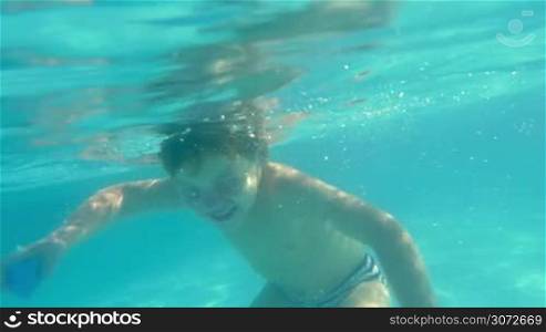 Slow motion shot of little boy swimming underwater in swimming pool and touching camera.