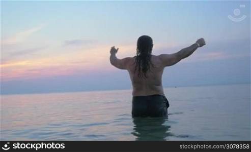 Slow motion shot of long-haired senior man stretching hands standing in the sea at sunset. Feeling free