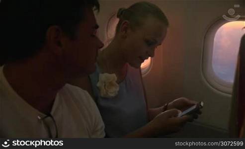 Slow motion shot of two people sitting on a plane. Woman is holding smartphone and shows something in it to her male friend.