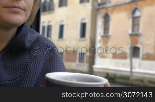 Slow motion, steadicam and close-up shot of a woman drinking hot tea from colorful cup sitting by the window at home