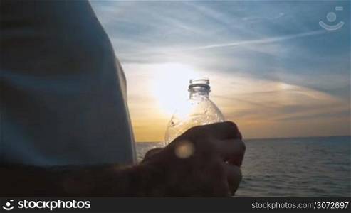 Slow motion, steadicam and close-up shot of a young man drinking water from plastic bottle by the sea at sunset. Water shining in the sun as he putting bottle down
