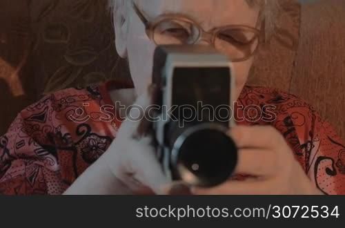 Slow motion, steadicam and close-up shot of an aged woman in glasses using retro video camera at home