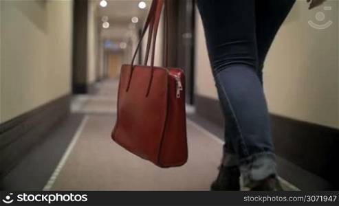 Slow motion steadicam and low angle shot of a woman in jeans with red leather bag walking along the light hotel corridor