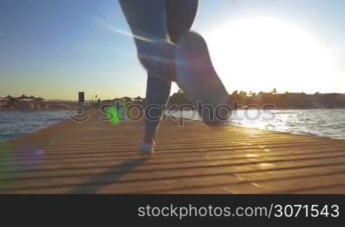 Slow motion steadicam and low angle shot of woman having evening training on resort. She running along the pier, bright sun shining and reflecting in the sea