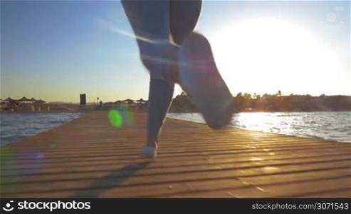 Slow motion steadicam and low angle shot of woman having evening training on resort. She running along the pier, bright sun shining and reflecting in the sea