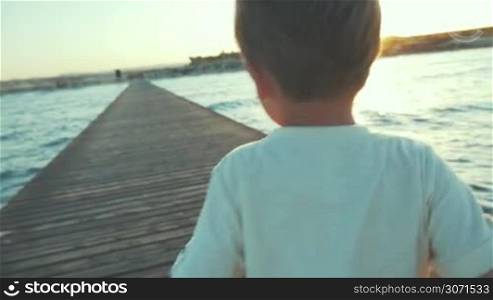 Slow motion steadicam back shot of a little boy running along the wooden pier in the sea at sunset