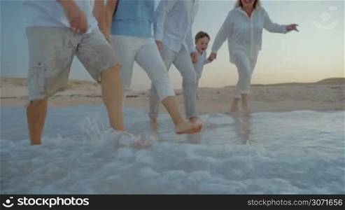 Slow motion steadicam, low angle shot of happy family with child having fun while splashing water with feet on the coast