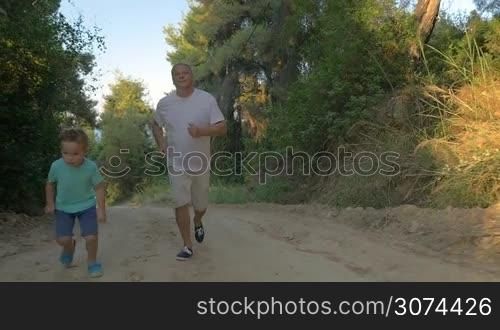 Slow motion steadicam shot of a child jogging with grandfather in green forest in the evening. Grandson is ahead of grandpa. Healthy outdoor activity