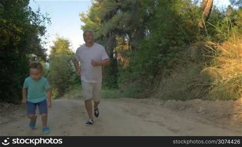 Slow motion steadicam shot of a child jogging with grandfather in green forest in the evening. Grandson is ahead of grandpa. Healthy outdoor activity