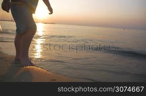 Slow motion steadicam shot of a little barefoot boy running on wet sand along the sea in the warm light of evening sun. Careless and happy childhood