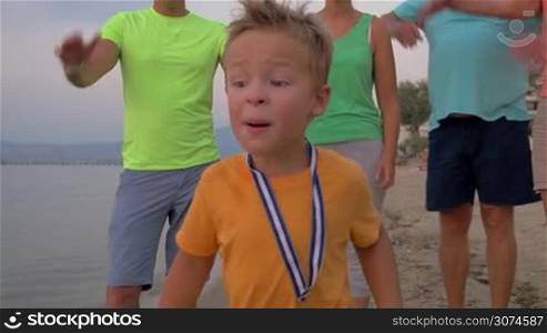 Slow motion steadicam shot of a little boy with medal and cup running along the beach, happy family applauding to his success. First victories