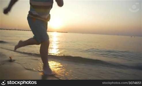 Slow motion steadicam shot of a little child running barefoot in water along the coast. Beautiful scene with sunset and endless sea in background