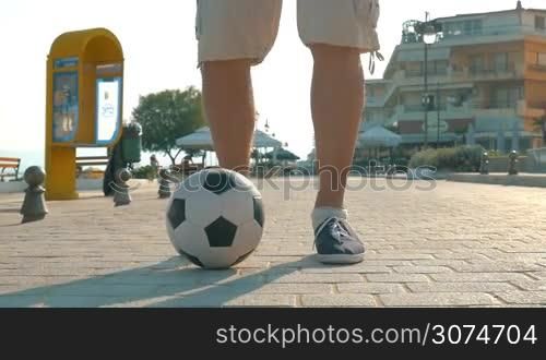 Slow motion steadicam shot of a man dribbling football along the sidewalk in resort town, only feet can be seen. Active life with sport