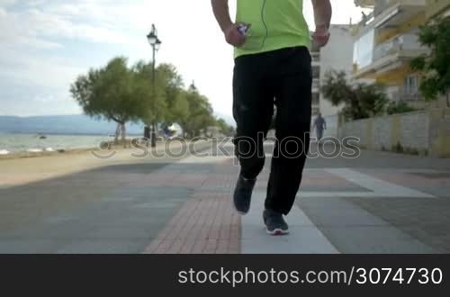 Slow motion steadicam shot of a man with smartphone jogging along the seafront in resort town. Active lifestyle and sports