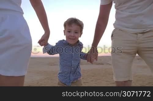 Slow motion steadicam shot of a playful little boy taking away parents hands and running away. Happy mother and father following him. Family fun on vacation