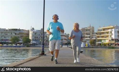 Slow motion steadicam shot of a senior man and woman in headphones running along the pier on resort. They listening to music with smart phones