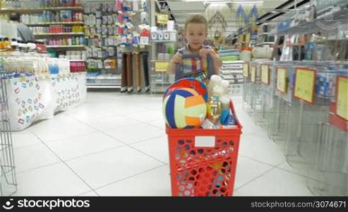 Slow motion steadicam shot of a smiling boy with full shopping cart moving towards the camera in the household goods section of the supermarket
