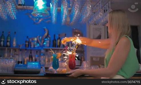 Slow motion steadicam shot of a young happy waitress setting on the Bengal fires in cocktails and carrying them through the bar