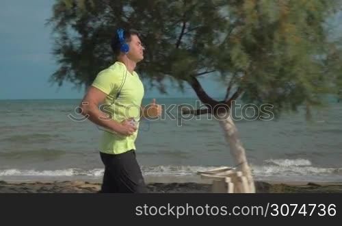 Slow motion steadicam shot of a young man having a run along empty beach. He listening to music in headphones and using smartphone
