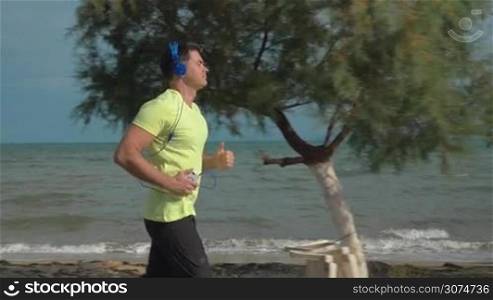 Slow motion steadicam shot of a young man having a run along empty beach. He listening to music in headphones and using smartphone