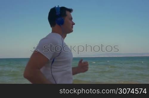 Slow motion steadicam shot of a young man having regular jog training along the coast, sea and clear blue sky in background. He listening to music in headphones and using smartphone while running