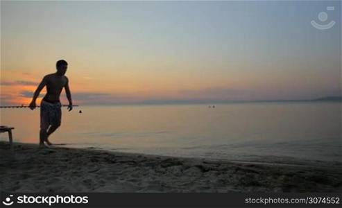Slow motion steadicam shot of a young man performing flips and somersaults on the beach at sunset. Acrobatics on the sand