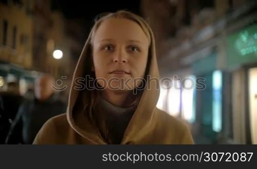 Slow motion steadicam shot of a young woman walking in the street late in the evening. She taking off the cowl, putting on the mask and continuing the way with happy smile