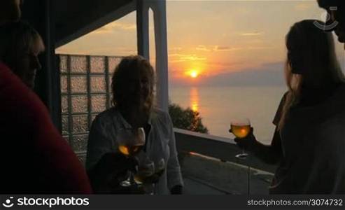 Slow motion steadicam shot of family clanging glasses on outdoor terrace of summer house at the seaside with scenic view to the sea and sunset