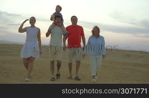 Slow motion steadicam shot of happy big family walking outdoor. Grandparents, young parents and little son riding on fathers neck enjoying evening on the beach