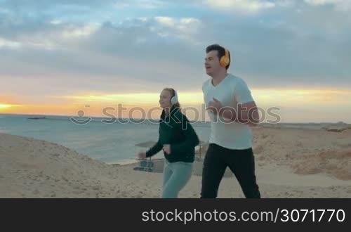Slow motion steadicam shot of happy couple running at the seaside at sunset. Enjoyable open air sports with music