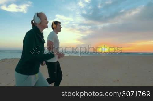 Slow motion steadicam shot of happy young people running along the beach on background of sunset over sea. Everyday training with music