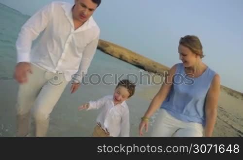 Slow motion steadicam shot of little boy and young parents having fun on the coast. They running barefoot in water and enjoying every moment of their happy vacation