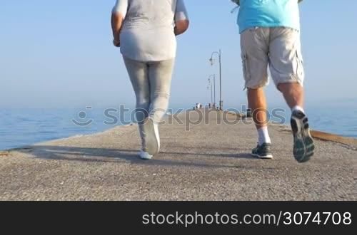 Slow motion steadicam shot of senior man and woman having a jog along the pier in the sea, back view. Active and healthy lifestyle