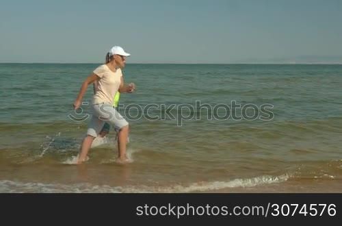 Slow motion steadicam shot of young happy man and woman running in sea and splashing water. Refreshing jogging on hot summer day