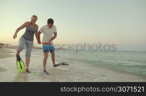 Slow motion steadicam shot of young man and woman walking in flippers along the sea shore. They look funny and clumsy